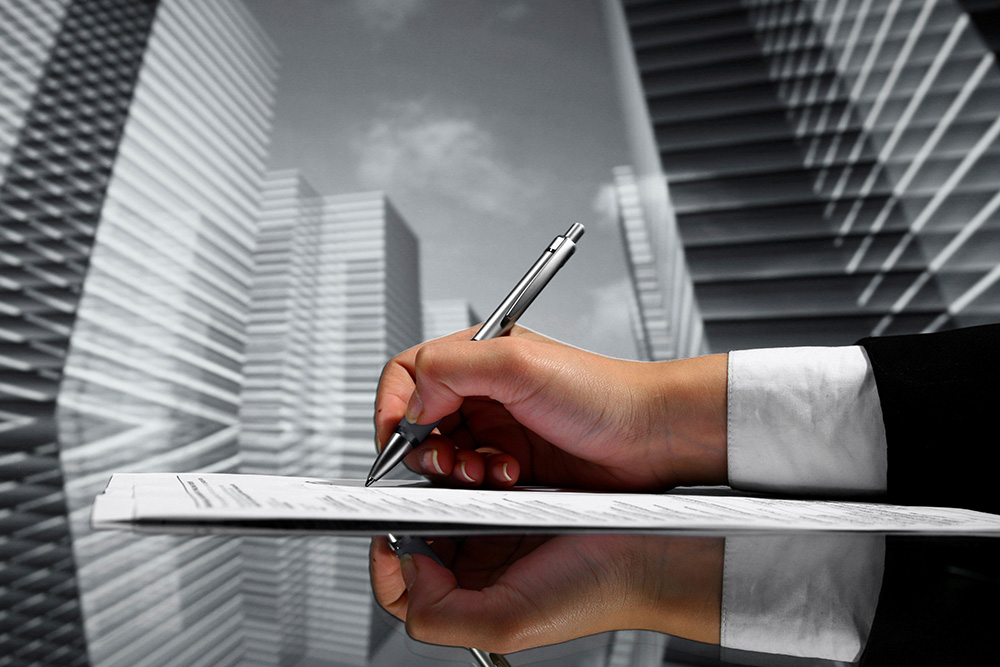 Image of a hand writing on a contract with skyscrapers in the background